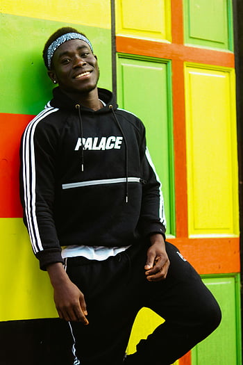 man leunt op de muur, Glimlachende jongeman, zwarte hoodie, levendige kleurrijke muur, buitenshuis, Paleis hoodie, Vrijetijdsoutfit, Street Style, stedelijke mode, hoofdband, zelfverzekerde pose