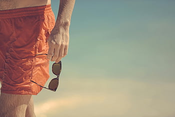 occhiali da sole con montatura nera, lente nera, persona in possesso di occhiali da sole, Shorts arancioni, spiaggia, stile estivo, look casual, scena all'aperto, Hand che tiene occhiali da sole, atmosfera di vacanza, Accessorio per occhiali da sole