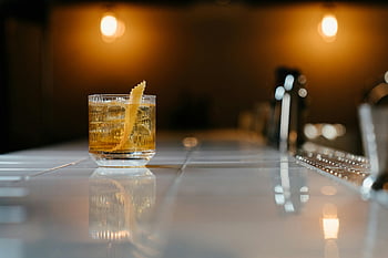 clear drinking glass, yellow liquid, whiskey cocktail, zest garnish, glossy bar counter, dimly lit setting, stylish drink, bar setting, cocktail glass, yellow cocktail, dim lighting