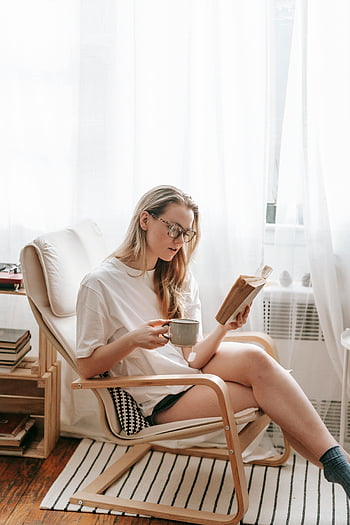 млада жена, четене на книга, фотьойл, Релаксираща сесия, У дома, напитка, комфортен, очила, Ежедневно облекло, Настройка на закрито, Уютна атмосфера