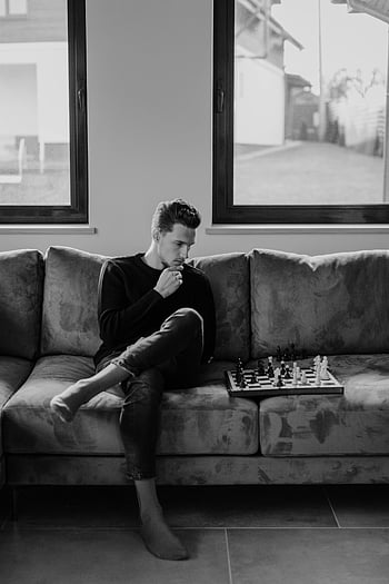 Man in Black Jacket, นั่งบนโซฟา, เล่นหมากรุก, ภาพขาวดำ, กำลังคิดจะเล่นหมากรุก, การตั้งค่าในร่ม, กระดานหมากรุก, ตัวหมากรุก, ชุดลำลอง, การแสดงออกที่ครุ่นคิด, ห้องนั่งเล่น