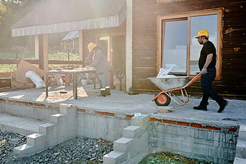 pracovníci na stavbě, Renovace domu, vnější práce, nesení materiálů, zpracování dřeva, Muž v černé košili, šedá a oranžová kolečka, exteriér domu, staveniště, Projekt renovace, venkovní práce