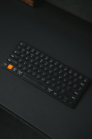 wireless keyboard, sleek black keyboard, orange button, tech setups, dark background, close-up, modern keyboard, compact keyboard, minimalist design, wireless technology, keyboard accessories