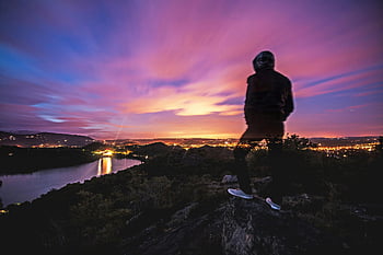man in black jacket, rocky ground, city lights, twilight, scenic overlook, long exposure, blurry figure, observing, sunset, landscape, nature