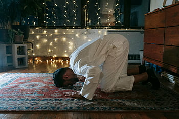 Muslim man, white thobe, praying, red and blue rug, traditional attire, indoor prayer, ambient lighting, kneeling, spiritual practice, cultural tradition, prayer rug