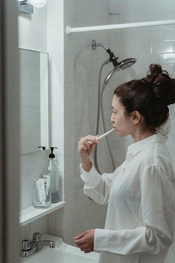 woman brushing teeth, modern bathroom, oral hygiene, toothbrush, bathroom mirror, daily routine, personal care, dental care, white shirt, shower head, sink