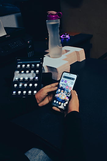机に座っている人、 スマートフォンを持っている、 マルチタスク、 現代技術の使用、 スマートフォンとタブレット、 机のセットアップ、 デジタル機器、 電話を使って手、 オフィス環境、 水筒、 ワークスペース