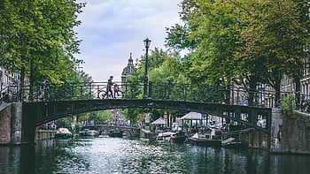 Amsterdam canal, classic bridge, bikes, trees, cloudy sky, person crossing bridge, picturesque view, Amsterdam, canal scene, urban landscape, European cityscape
