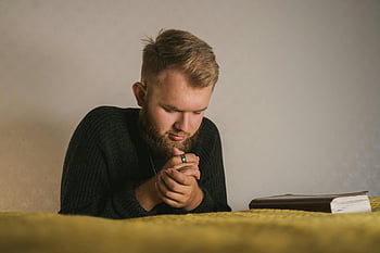 hombre barbudo, suéter de punto negro, rezando, Biblia, entorno interior sereno, fotografía de cerca, oración interior, momento espiritual, escena religiosa, ambiente tranquilo, reflexión personal