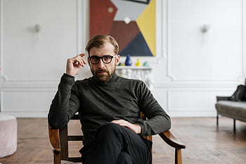 Muž v černém svetru, hnědá dřevěná židle, moderní interiér, abstraktní umění, muž s brýlemi, Sedící muž, Současná místnost, Stylový muž, Vnitřní nastavení, příležitostná pozice, umělecké zázemí