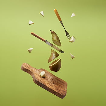 sliced pear, suspended midair, fresh pear slices, kitchen utensils, zero gravity effect, light green backdrop, floating pear, wooden cutting board, knife, fork, fruit photography