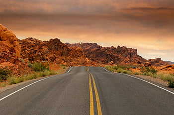 scenic highway, red desert rock, dramatic rock formations, colorful sunset sky, golden hour, gray concrete road, brown mountain, desert landscape, winding road, natural beauty, outdoor scenery