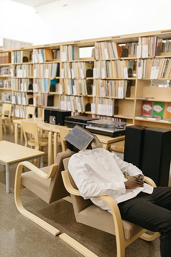 man sleeping, book on face, library, student, modern library, bookshelves, study area, comfortable chair, quiet environment, educational setting, reading space