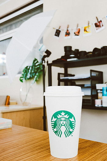 Starbucks coffee cup, white disposable cup, brown table, wooden table, photography studio, stylish setting, indoor scene, green logo, studio lighting, coffee cup, studio props