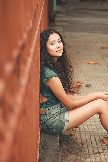 žena sedící, kudrnaté vlasy, venkovní portrét, Neformální outfit, u zdi, Mladá žena, džínové šortky, zelený top, přirozené světlo, městském prostředí, pouliční fotografie