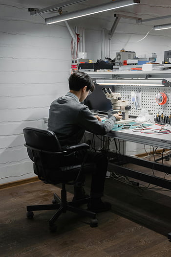 man sitting, electronics workspace, engineer focusing, project work, well-equipped lab, microscope, desk setup, workspace tools, technical environment, indoor workspace, professional setting