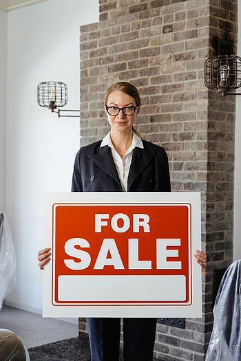 real estate agent, female real estate agent, for sale sign, holding sign, ready to assist, clients, indoor setting, brick wall, professional attire, real estate marketing, property sale