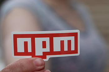 npm sticker, red npm, hand holding, web development, close-up photo, blurred background, development themes, npm logo, person holding, red sticker, web design
