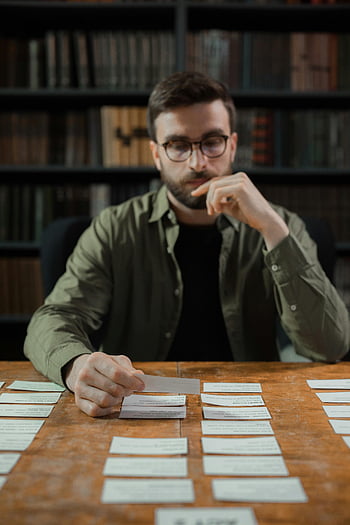 bearded man, eyeglasses, sorting documents, library setting, pieces of paper, table, concentration, man with glasses, document organization, study environment, bookshelves