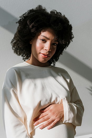 zwangere vrouw, witte jurk met lange mouwen, Natuurlijk daglicht, Serene expressie, Buik omarmen, zachte verlichting, afro haar, zwangerschapsmode, zwangerschapsstijl, natuurlijke schoonheid, rustige sfeer