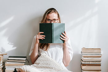 young woman, eyeglasses, reading book, indoor, surrounded by books, calm atmosphere, thoughtful, homework assignment, stacks of books, serious, indoor reading