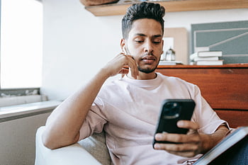 indisk mand, trådløse øretelefoner, Messaging -smartphone, tankevækkende udtryk, ung mand, indendørs indstilling, fokuseret look, komfortable siddepladser, Brug af smartphone, afslappet tøj, indendørs miljø