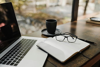 černé orámované brýle, poznámkový blok, Otevřený notebook, káva, dřevěný stůl, Útulný pracovní prostor, Brýle na poznámkovém bloku, nastavení pracovního prostoru, stolní příslušenství, Nástroje produktivity, kávový šálek