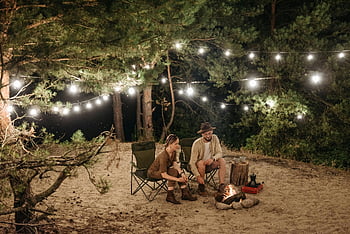 Paar Camping, Lagerfeuer, Lichterketten, Waldeinstellung, Nachtcamping, Erholung im Freien, Campingstühle, Romantisches Camping, Naturcamping, Gemütliches Camping, Lagerfeuer kochen