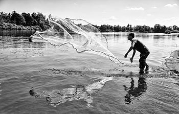 grayscale photo, man throwing fishing net, black and white, fisherman casting net, river reflections, serene scene, outdoor fishing, water activity, traditional fishing, nature photography, monochrome image