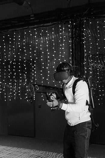 grayscale photo, man playing, virtual reality game, VR gun, monochrome setting, virtual reality simulation, man engaged, VR headset, string lights, indoor setting, gaming experience