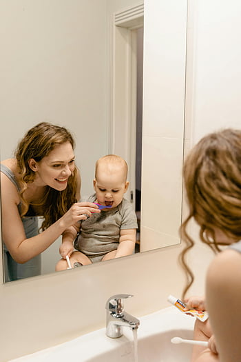 Matka kartáčování dětských zubů, Domácí koupelna, Hygienové návyky, dětský zubní kartáček, koupelnové zrcadlo, matka a dítě, Péče o zuby, zubní pasta, dřez, kohoutek, Rodinná hygiena