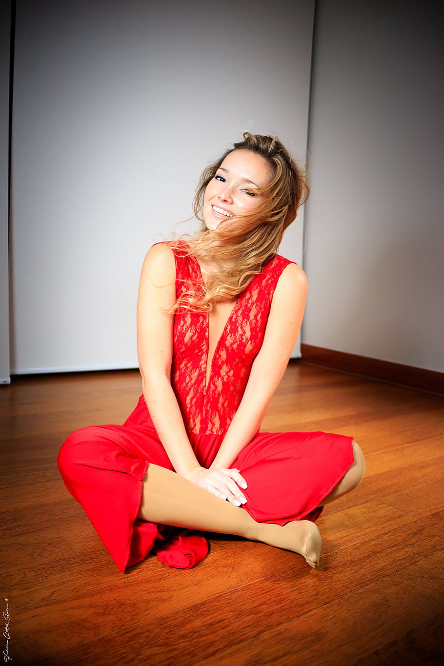 red dress, sitting pose, wooden floor, blonde hair, studio lighting, casual style, indoor setting, smiling woman, fashion photography, relaxed posture, vibrant color