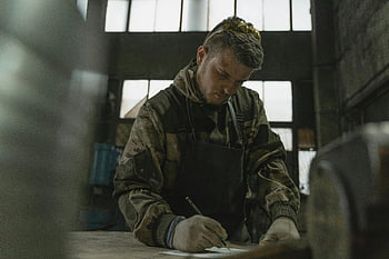 man drawing, paper sketching, dimly lit, industrial factory, camouflage attire, man sketching, industrial setting, drawing paper, man in camouflage, factory environment, sketching man