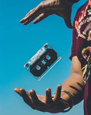 retro cassette tape, floating cassette, hands holding cassette, clear blue sky, evoking nostalgia, vintage music, cassette tape tricks, outdoor cassette, nostalgic cassette, floating object, retro music