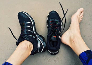 Black Under Armour sneakers, bare feet, gray surface, athletic shoes, Under Armour, sports footwear, black sneakers, casual shoes, footwear, athletic wear, running shoes