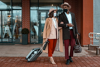 おしゃれなカップル、 フェイスマスク、 ホテルの入り口、 パンデミック中の旅行、 コートを着ている女性、 スーツを着た男、 荷物、 れんが造りの建物、 屋外のシーン、 スタイリッシュな衣装、 パンデミックの安全