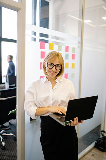 woman with eyeglasses, holding laptop, smiling businesswoman, modern office setting, glass walls, sticky notes, professional woman, office environment, business attire, technology in office, workplace productivity