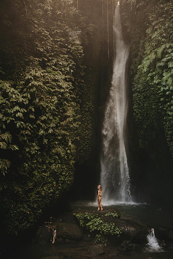 woman in bikini, standing on rock, near waterfall, tropical serenity, lush jungle, Bali, majestic waterfall, base of waterfall, natural beauty, travel photography, adventure travel