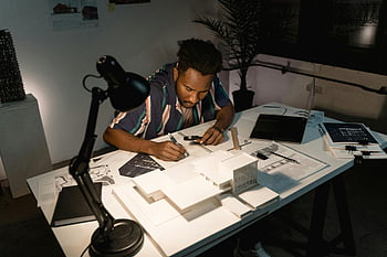 man writing notes, work table, focused architect, desk lamp, sketching designs, models, blueprints, architect working, desk, design models, blueprints under lamp