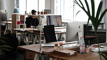 Mann sitzt, Computer, Open-Plan-Büro, moderne Einrichtung, natürliche Beleuchtung, Produktivität, Schreibtisch, Arbeitsplatz, Zimmerpflanzen, Büroumgebung, arbeitsfrei