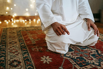 man in white thobe, sitting, red and brown area rug, traditional attire, kneeling in prayer, indoors, string lights overhead, cultural clothing, prayer position, indoor setting, string lights