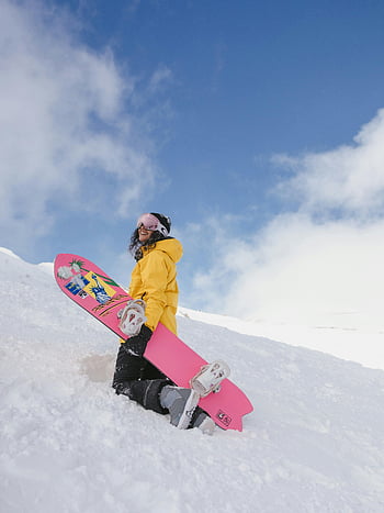 Γυναίκα σε κίτρινο hoodie, ροζ snowboard, χιονισμένο βουνό, γαλάζιος ουρανός, χειμωνιάτικη ενδυμασία, χαμογελαστή γυναίκα, χιονοσανίδα, υπαίθρια χειμερινά σπορ, κλίση που καλύπτεται από χιόνι, χειμερινή αναψυχή, ροζ και λευκό snowboard