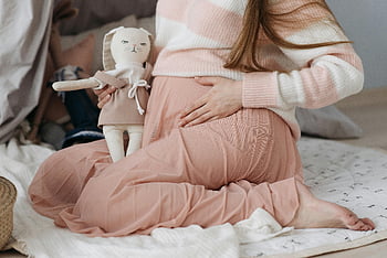 raskaana oleva nainen, vaaleanpunainen hame, Kosketa vatsaa, odottava äiti, istuu lattialla, pehmeä lelu, pinkki ja valkoinen asuste, kodikas vaatteet, äitiysmuotia, raskauden tyyli, vauva kolahtaa