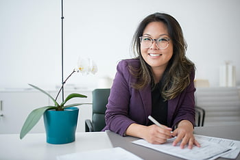 Aziatische vrouw, bril, glimlachen, werkend, bureau, documenten, plant in bloempot, kantooromgeving, professionele kleding, binnen-, werkruimte
