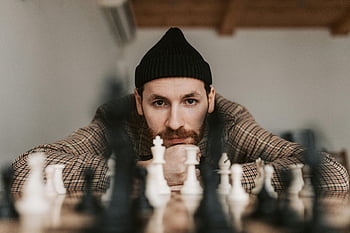 Muž na sobě černou beanie, šachová hra, soustředěný muž, Vnitřní nastavení, Promyšlená nálada, intenzivní výraz, strategické šachy, čepice, šachové figurky, Vnitřní šachová hra, Muž v Beanie