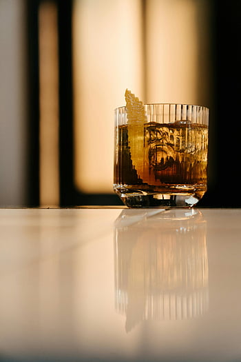 clear drinking glass, brown liquid, stylish cocktail, lemon zest garnish, reflective bar counter, classy evening, bar drink, cocktail glass, elegant drink, bar setting, brown cocktail
