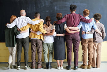 group of people, standing indoors, diverse group, adults, casual outfits, hugging, chalkboard, teamwork, indoor setting, casual attire, group hug