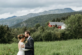 wedding couple, intimate moment, scenic mountain setting, man in suit jacket, woman in wedding dress, romantic scene, outdoor wedding, mountain backdrop, rural landscape, red barn, green field