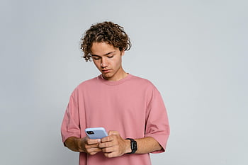 صبي مراهق،  تي شيرت وردي،  الهاتف الذكي الأزرق،  خلفية واضحة،  باستخدام الهاتف الذكي،  الزي عارضة،  شعر مجعد،  ساعة اليد،  إعداد داخلي،  تعبير مركّز،  استخدام التكنولوجيا
