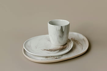 ceramic flatware, marble-patterned cup, minimalistic design, neutral background, ceramic plates, close-up photo, modern tableware, white ceramic, elegant dinnerware, simple aesthetic, flatware set